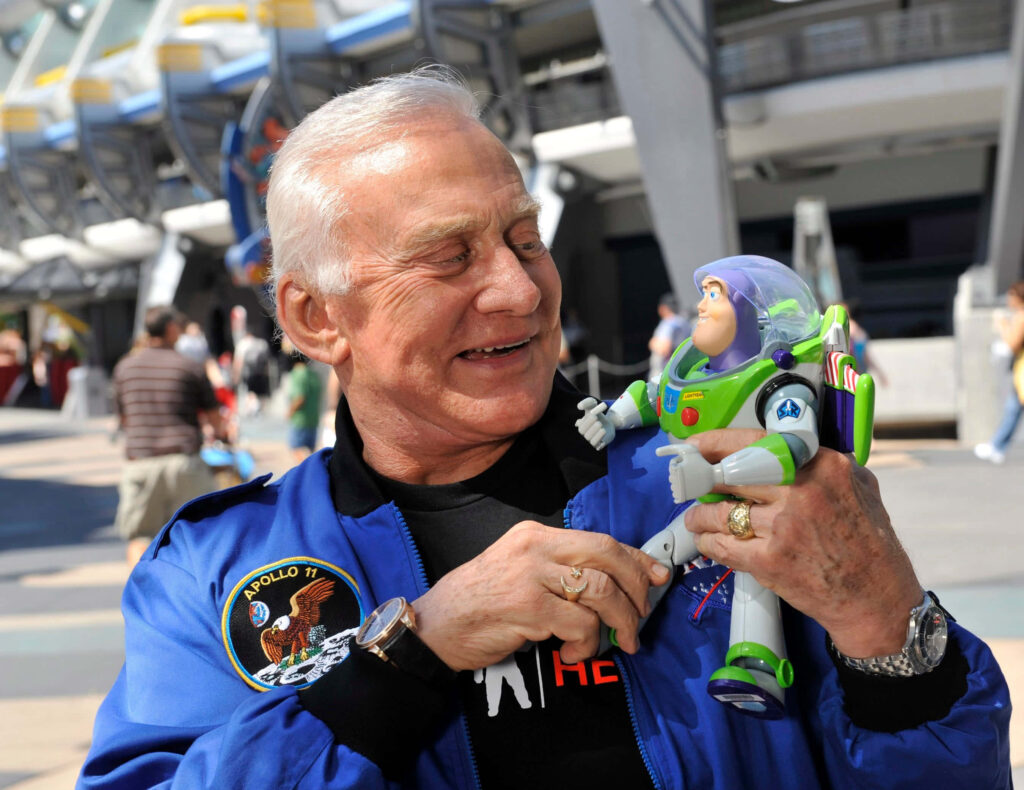 Buzz Aldrin in blue Apollo 11 jacket smiling and holding Buzz Lightyear toy outdoors vibrant scene Wallpaper