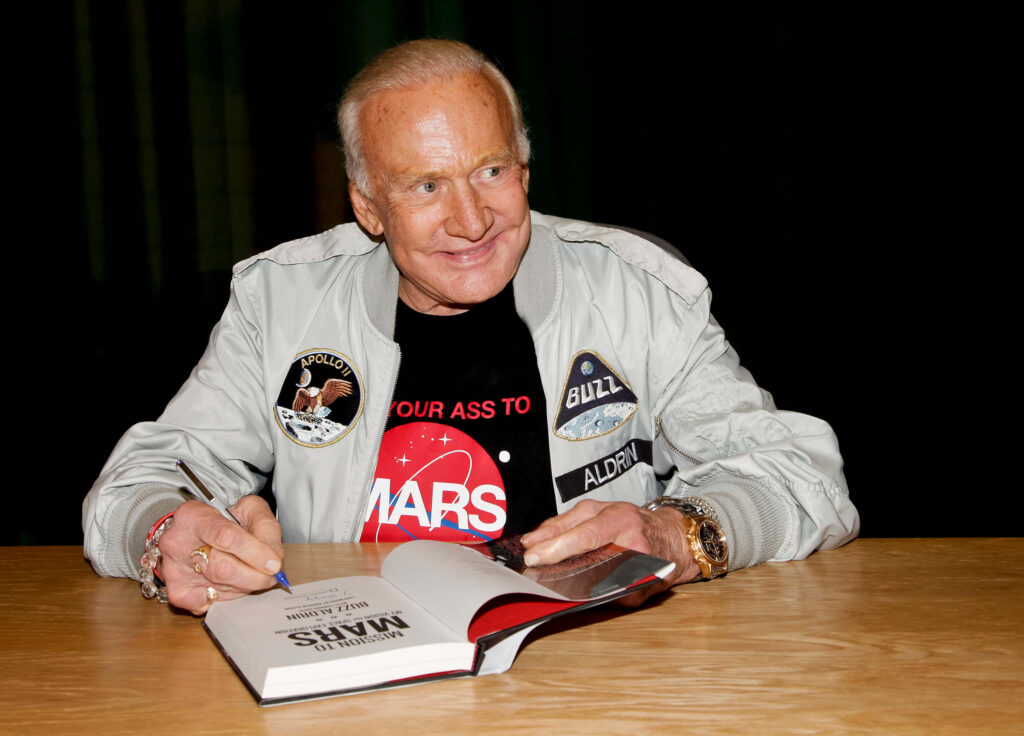 Buzz Aldrin signing autographs in NASA jacket with Apollo patches and Mars shirt vibrant wallpaper