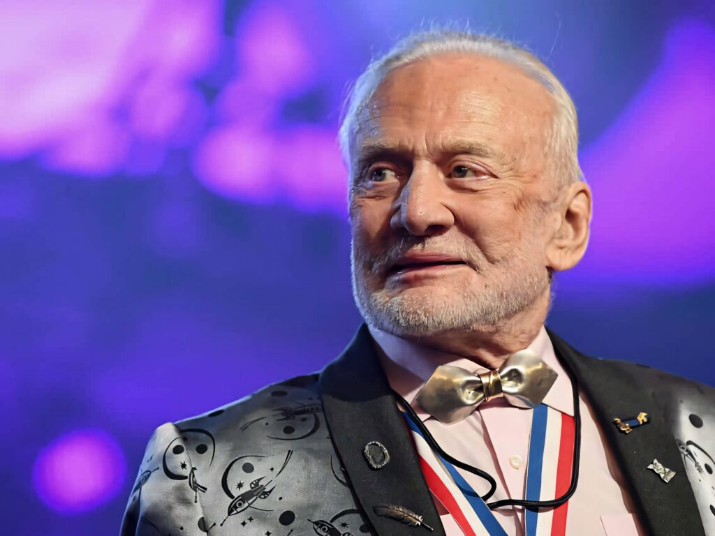 Buzz Aldrin at space-themed event wearing jacket with space motifs vibrant purple blue background Wallpaper