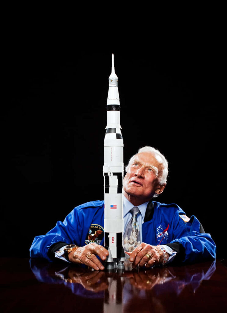 Buzz Aldrin in blue astronaut jacket with Saturn V rocket model cosmic exploration portrait Wallpaper