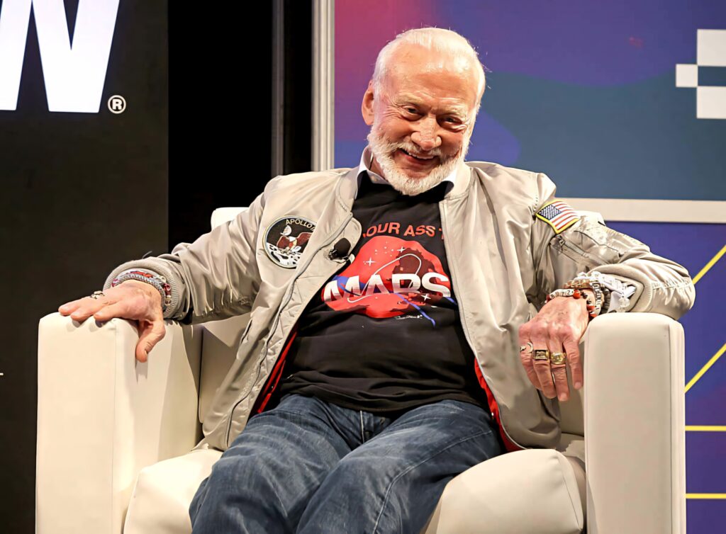 Buzz Aldrin in metallic NASA jacket and Mars T-shirt sitting in modern chair vibrant space wallpaper