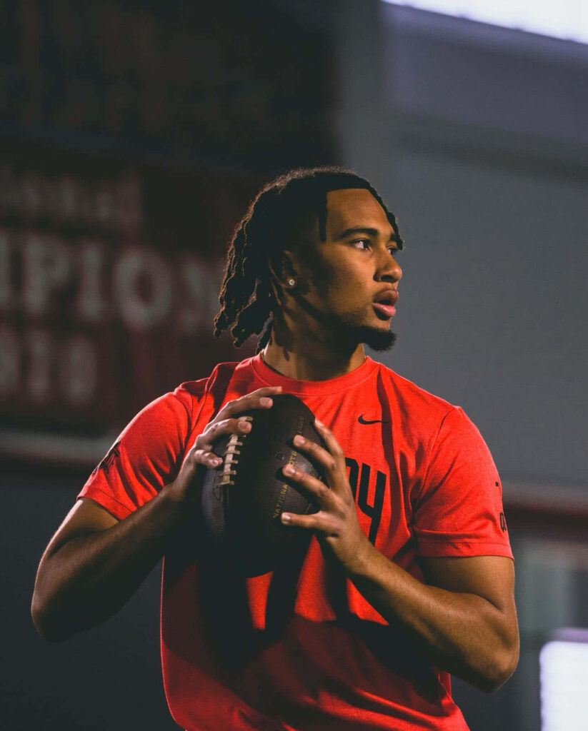CJ Stroud focused holding football in red shirt with dramatic lighting and blurred stadium background Wallpaper