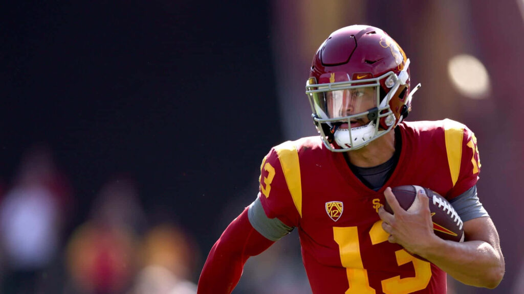 Caleb Williams in USC uniform throwing football with focus in blurred stadium action background Wallpaper