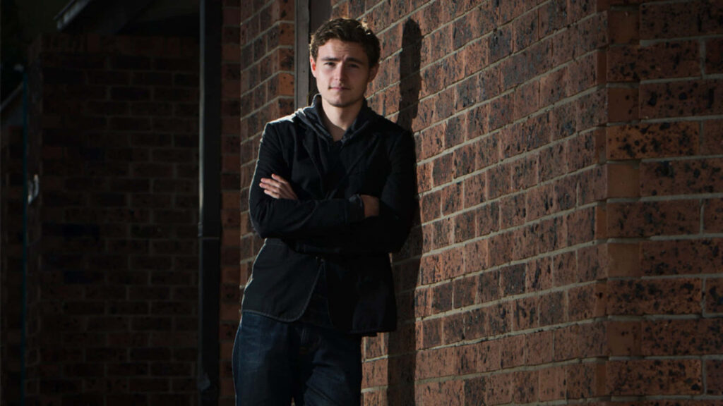 Callan McAuliffe leaning on brick wall wearing dark jacket jeans confident moody lighting wallpaper