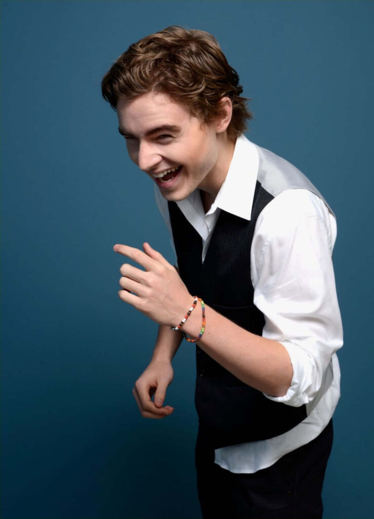 Callan McAuliffe playful portrait blue background white shirt black vest colorful beaded bracelet Wallpaper