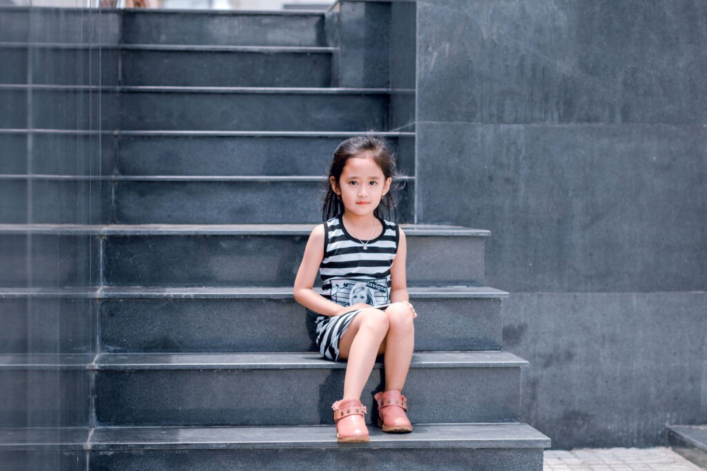 Young girl in striped dress sitting on dark stone steps against minimalist grey wall background Wallpaper
