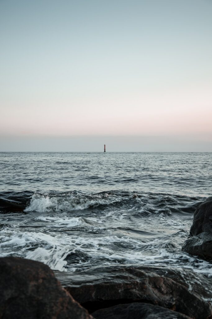 Calm coastal wallpaper with gentle sea waves dark shore rocks pastel sky and distant red lighthouse