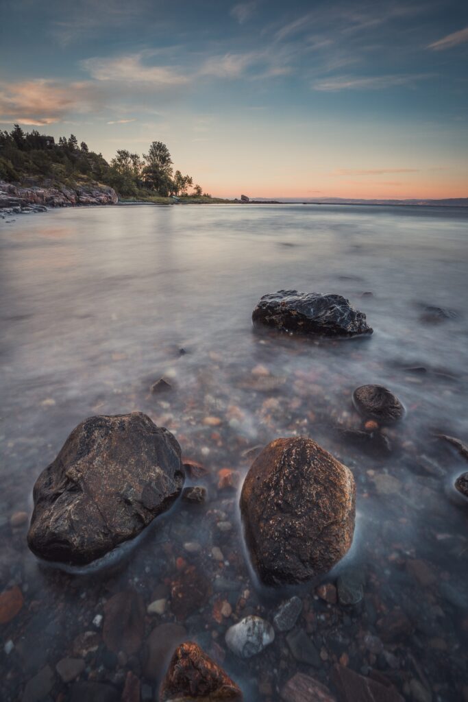 Calm lakeside with smooth stones clear water pastel sunset sky and distant tree horizon reflection Wallpaper