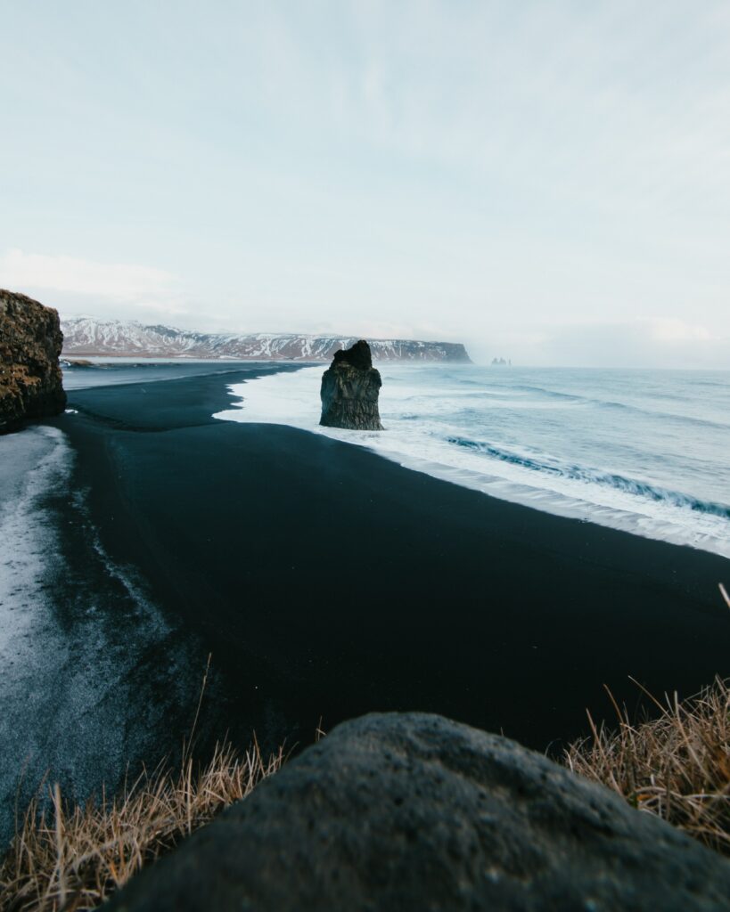 Rugged coastal rocks on black sand with calm ocean waves under an overcast sky wallpaper