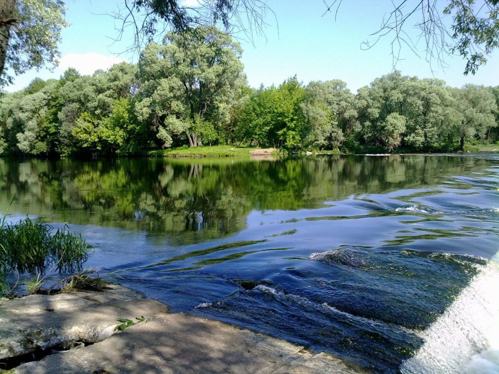 Calm river flowing over stone weir with green trees reflecting on blue water under sun Wallpaper