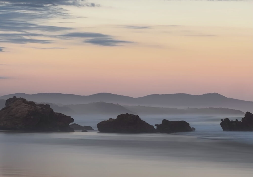 Rugged rocks in calm sea at dawn with misty fog and pastel orange blue sky wallpaper background