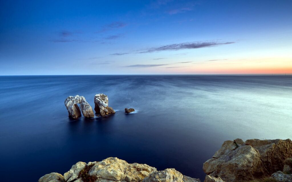 Calm sea with smooth glass water and rugged stone formations under clear blue sky wallpaper