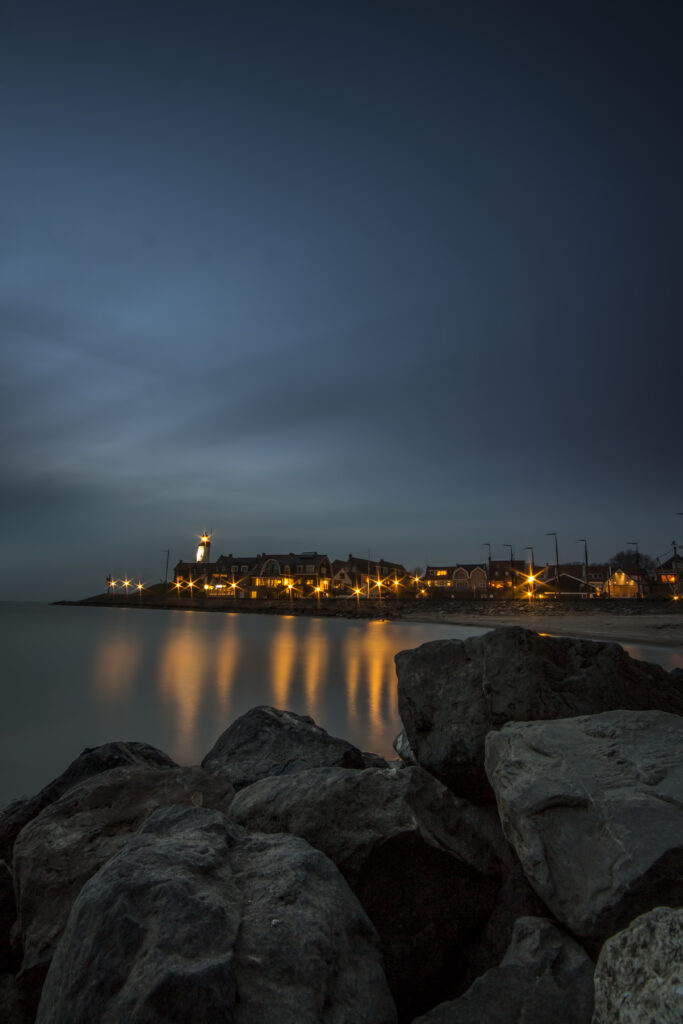 Dusk seaside with large stones warm lit houses lighthouse golden reflections calm water wallpaper