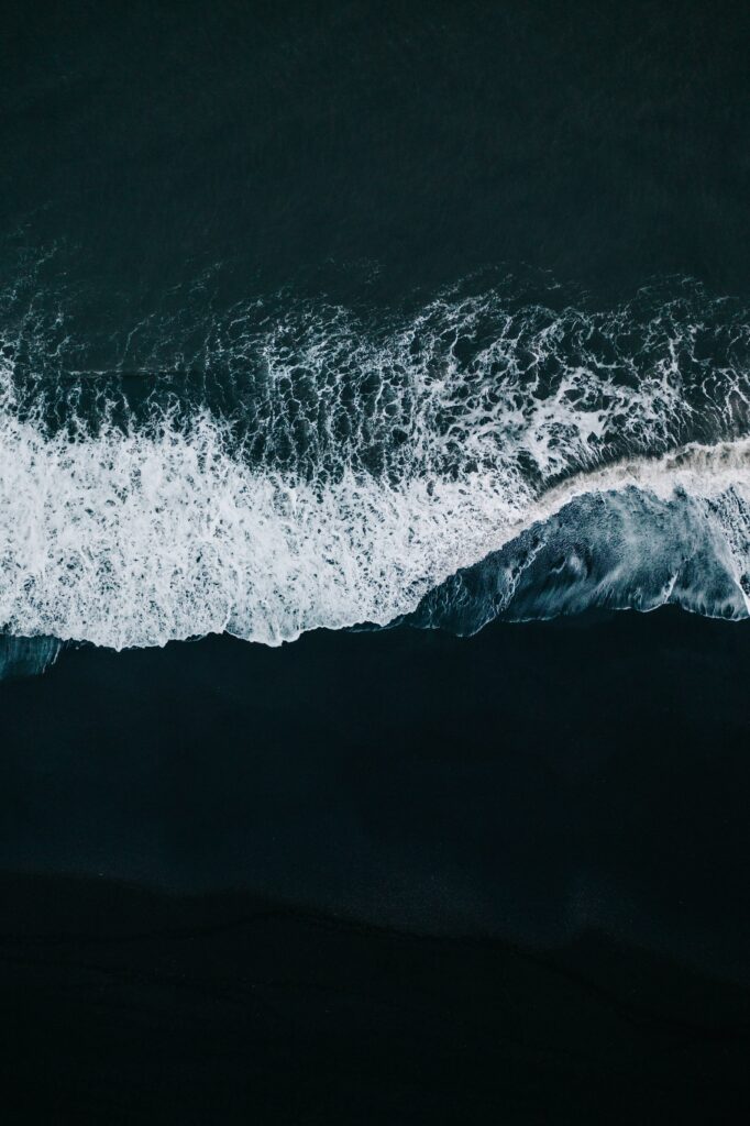 Aerial view of ocean waves crashing on dark sandy beach with vibrant sea foam and deep blue water Wallpaper