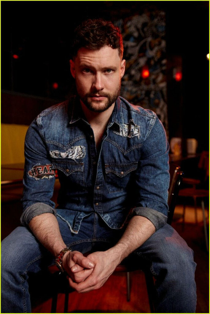 Calum Scott in denim jacket and jeans with warm lighting in modern dimly lit wallpaper background