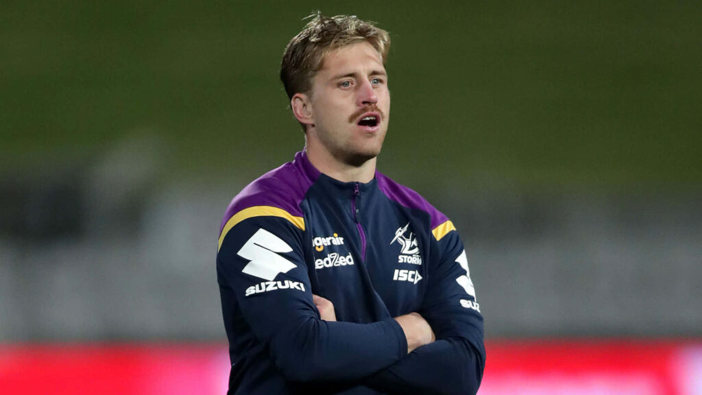 Cameron Munster in Melbourne Storm jacket with folded arms focused in blurred stadium background Wallpaper