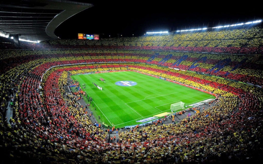 Camp Nou Stadium glowing at night with vibrant red and yellow crowd cheering during football match Wallpaper