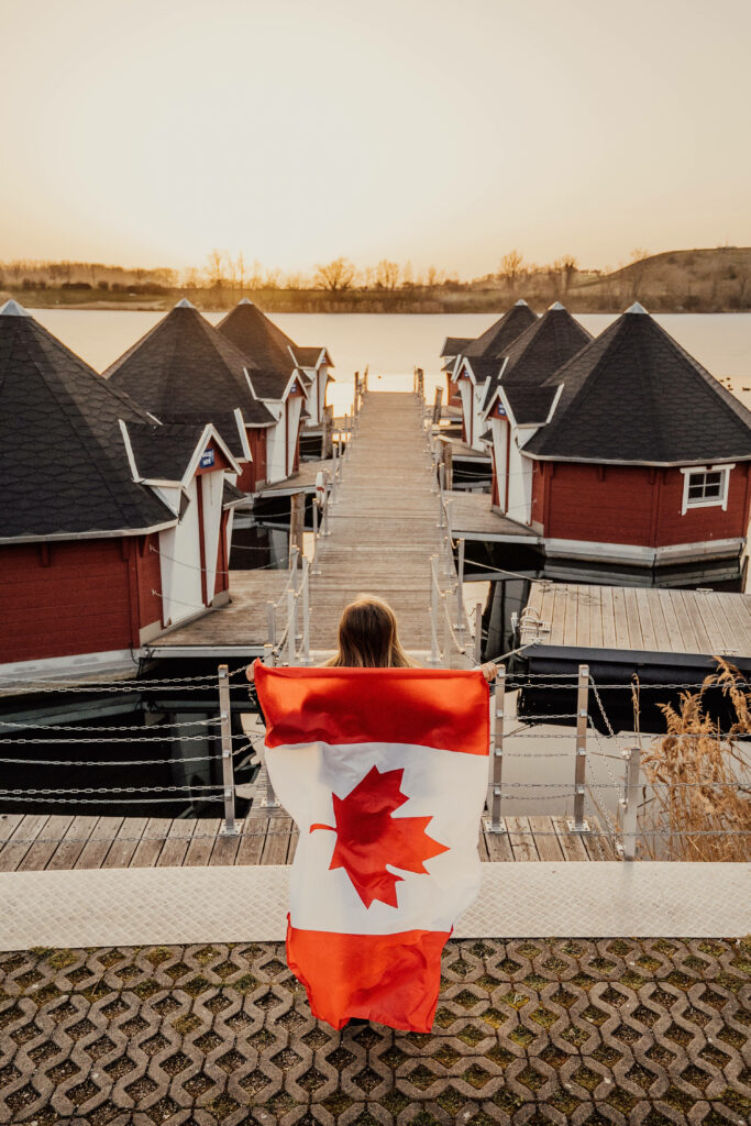 Woman with Canadian flag overlooking peaceful lakeside village of red cottages at sunset Wallpaper