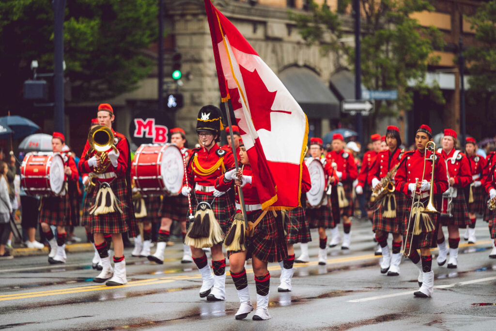 Canadian marching band in red uniforms and tartan kilts parading with flag on city street wallpaper