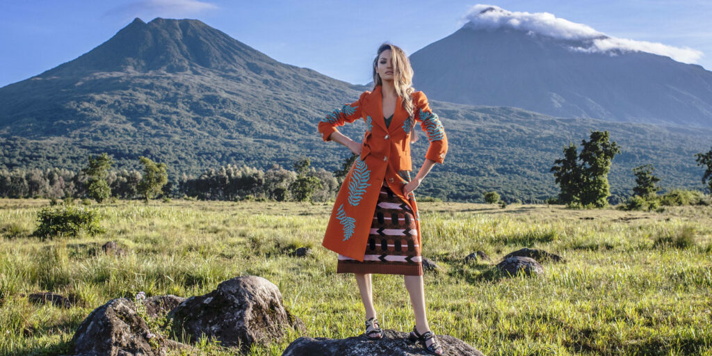 Candice Swanepoel in vibrant attire in Rwanda grasslands with volcanic mountains and blue sky background Wallpaper