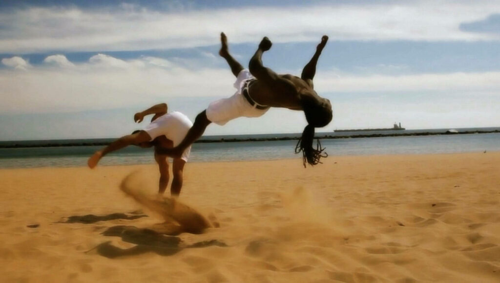 Two capoeira practitioners performing dynamic high kicks on sandy beach with sea background Wallpaper