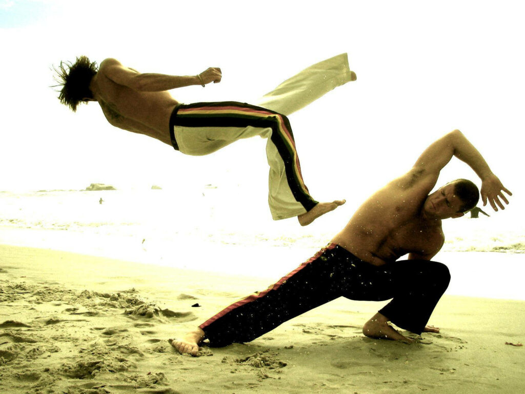 Two capoeira fighters perform acrobatic martial arts moves on a sunny sandy beach background Wallpaper