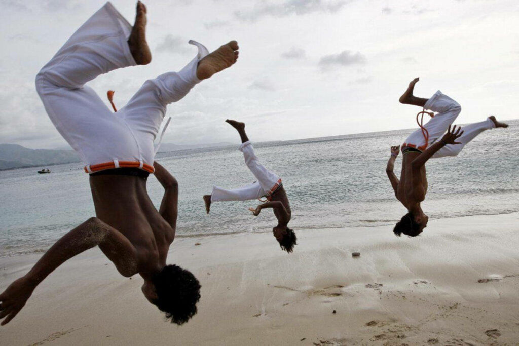 Three capoeira fighters perform acrobatic flips on a sandy beach with an ocean backdrop Wallpaper