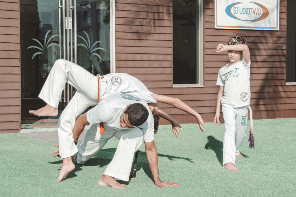 Capoeira practitioners performing dynamic somersault outdoors on grass while youth observes learning Wallpaper