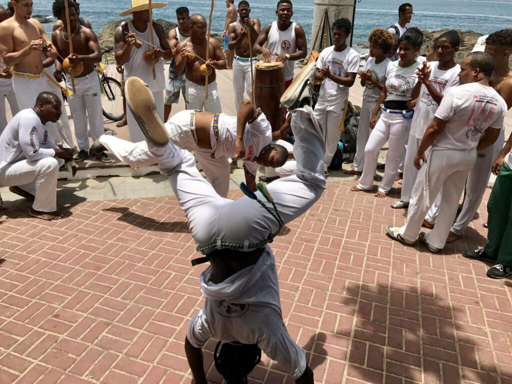 Two capoeira practitioners spar on brick walkway by ocean surrounded by musicians and spectators Wallpaper