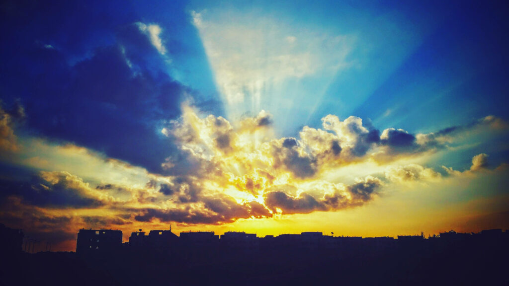 Sunset with golden rays over silhouetted city skyline and deep blue textured clouds wallpaper