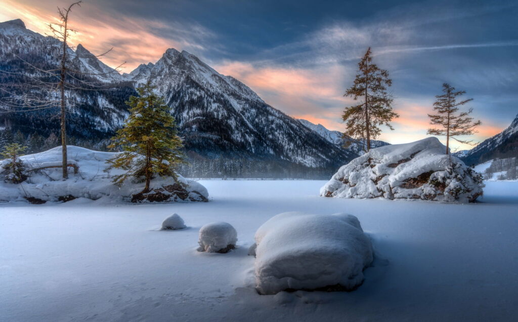 Snow-covered frozen Hintersee lake with Bavarian Alps mountains and pine-topped rocks at sunrise Wallpaper