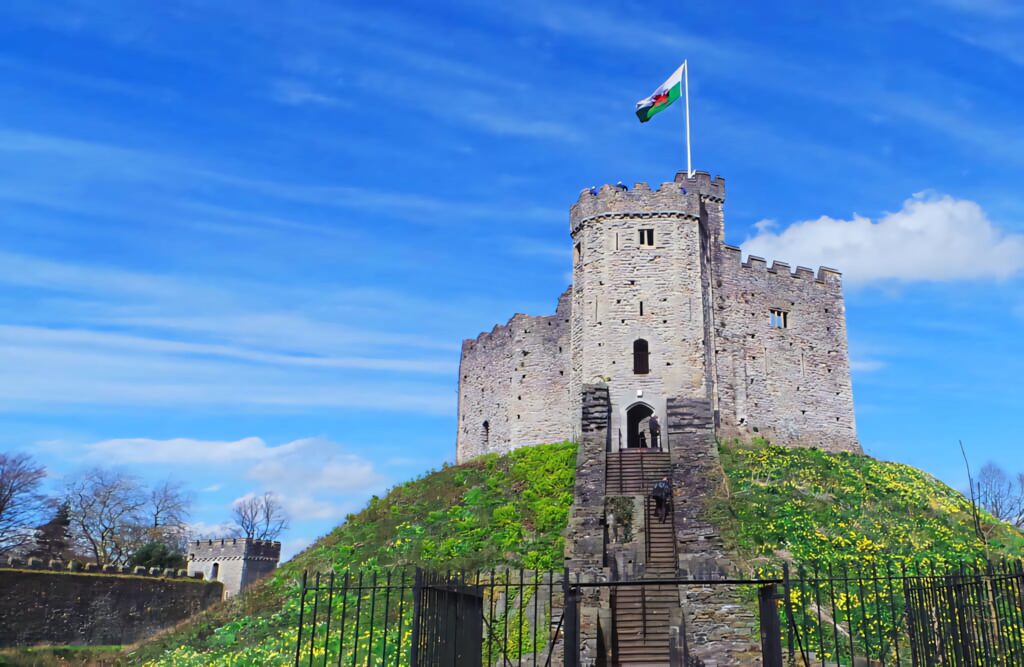 Cardiff Castle on grassy hill with detailed battlements Welsh flag bright blue sky wallpaper background