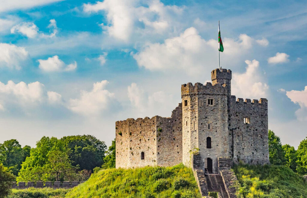 Cardiff Castle on green hill surrounded by trees under vibrant blue sky with soft clouds wallpaper