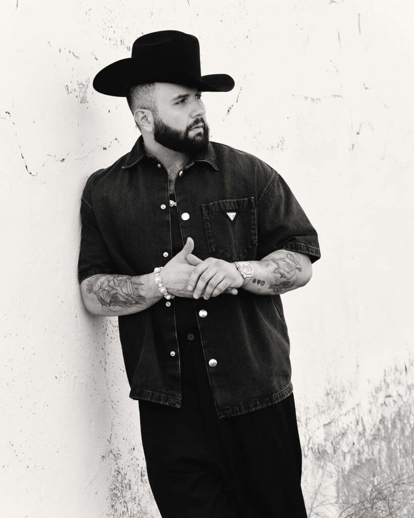 Carin León black and white wallpaper with tattoos wearing black hat shirt leaning on wall