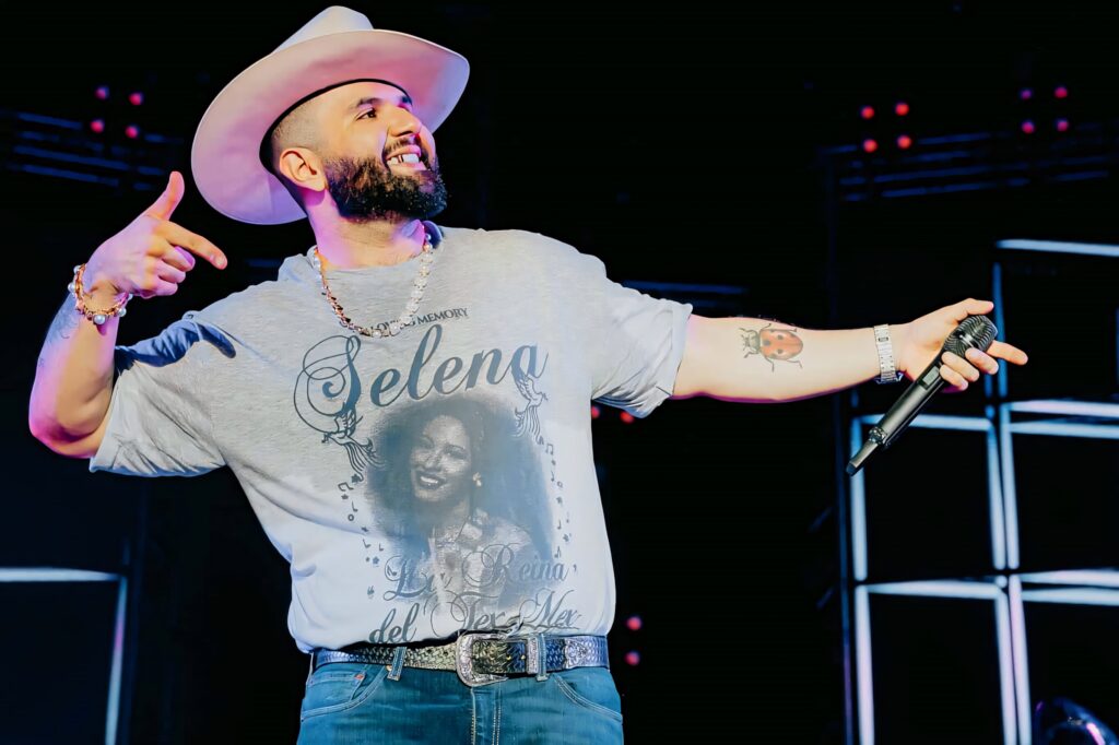 Carin León performing on stage wearing Selena tribute shirt and cowboy hat with vibrant lighting Wallpaper