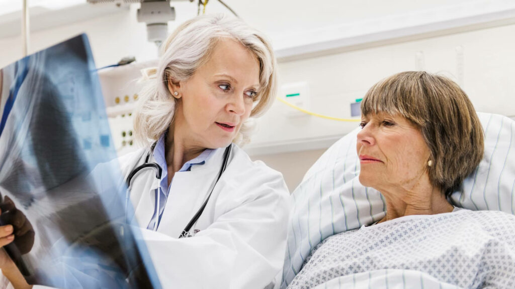 Doctor with gray hair explains X-ray to patient in hospital bed showing trusted medical care Wallpaper