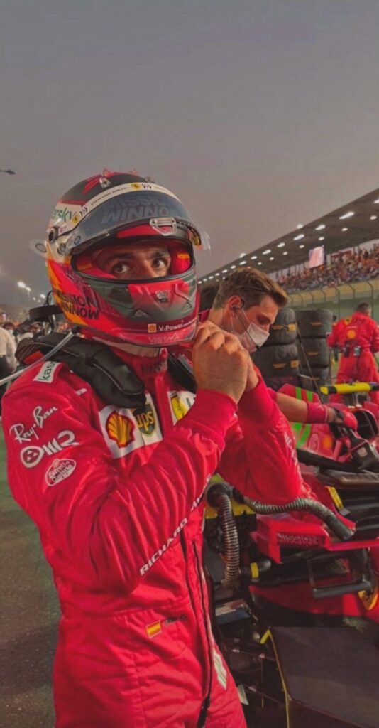 Carlos Sainz Jr. removing helmet in red Ferrari suit with vibrant racetrack and pit lane background Wallpaper