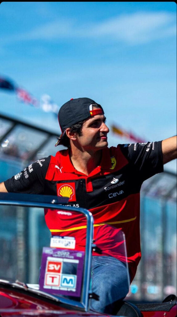 Carlos Sainz Jr in Ferrari racing gear waving to fans during vibrant Grand Prix motorcade event Wallpaper