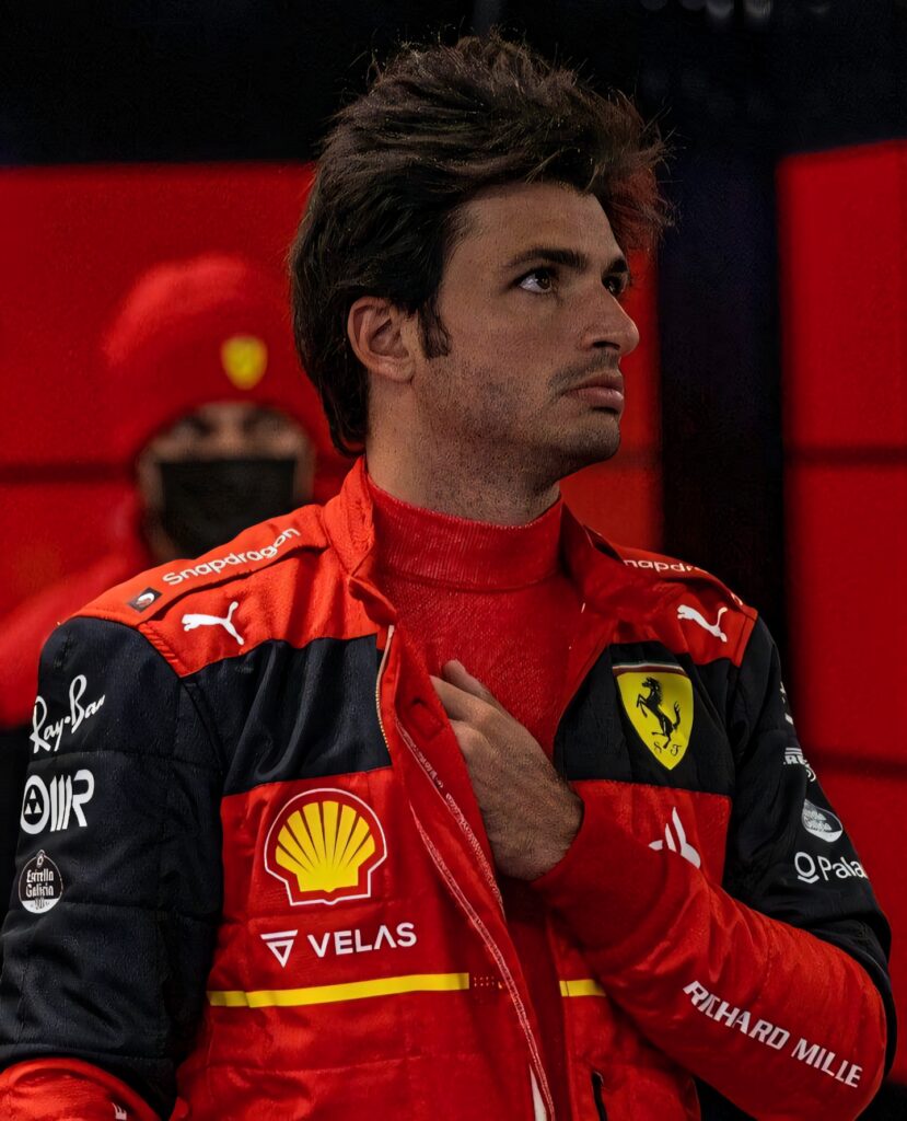 Carlos Sainz Jr in red Ferrari suit focused in pit area during high stakes car race wallpaper background