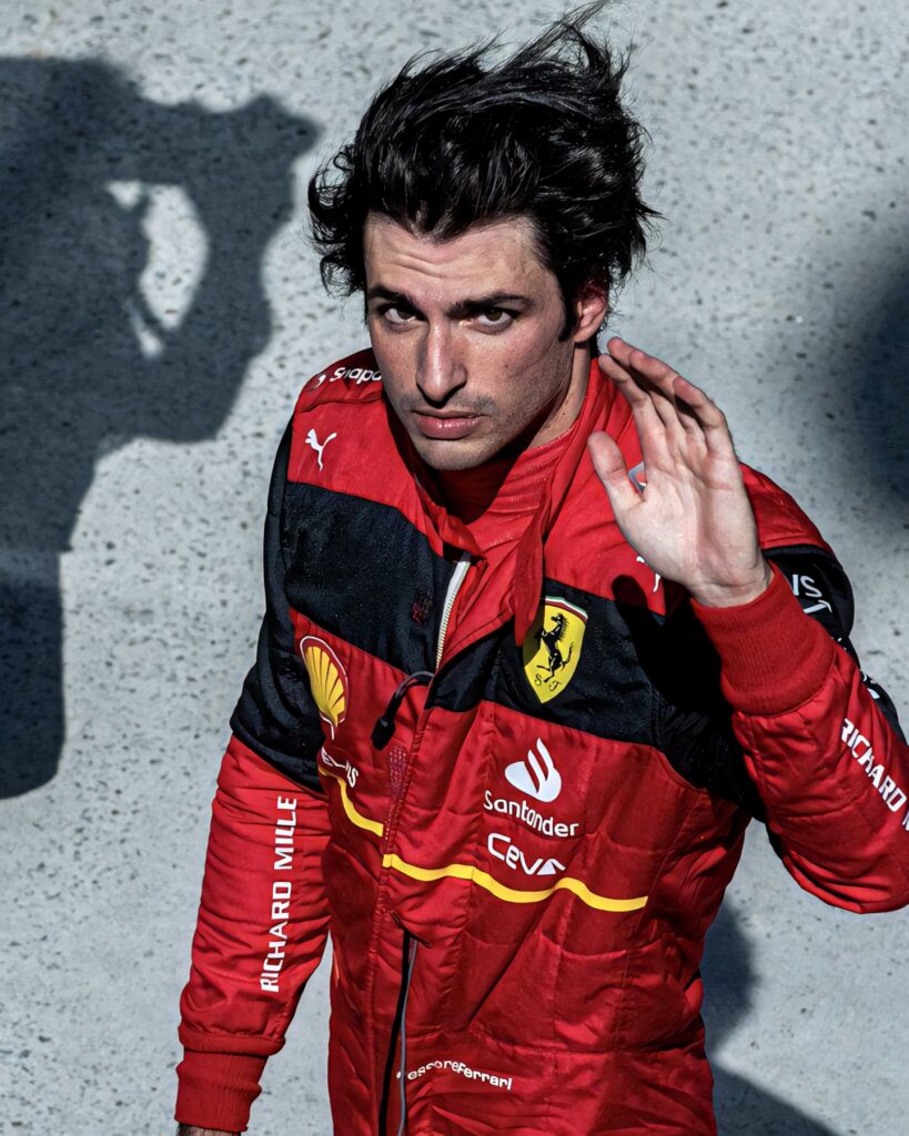 Carlos Sainz Jr in red Ferrari racing suit waving to fans with vivid sponsor logos and focused look Wallpaper