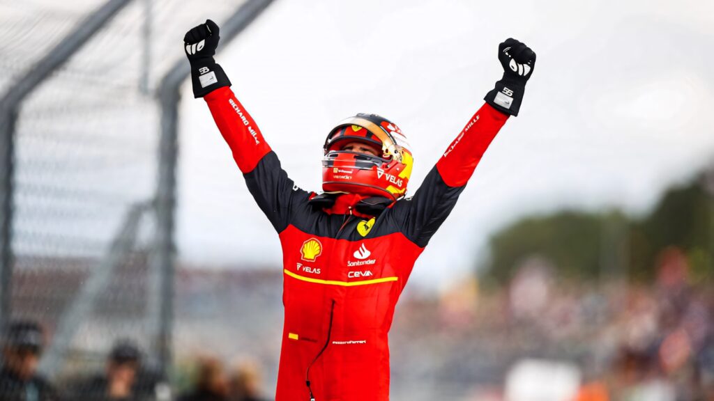 Carlos Sainz Jr in red Ferrari suit celebrating victory with arms raised on racetrack background Wallpaper
