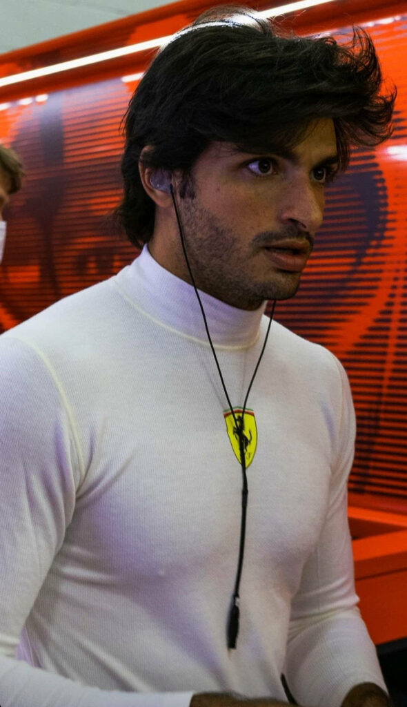 Carlos Sainz Jr focused in white Ferrari racing suit with earphones vibrant red Formula 1 paddock background Wallpaper