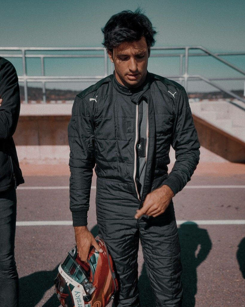 Carlos Sainz Jr in black racing suit holding helmet on sunlit racetrack focused motorsport portrait Wallpaper