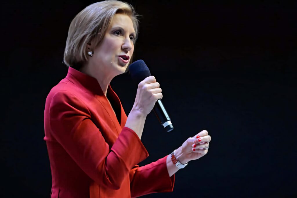 Carly Fiorina speaking passionately in red blazer with dynamic hand gesture on dark stage Wallpaper