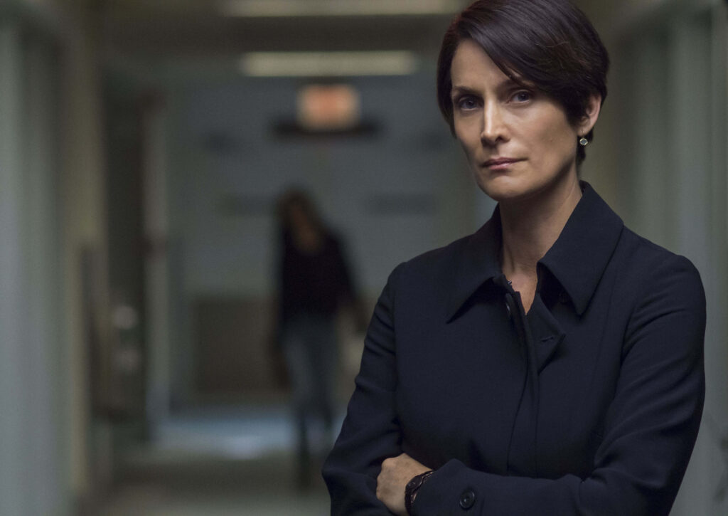 Carrie-Anne Moss confident pose in dim hallway with dramatic lighting and muted tones wallpaper