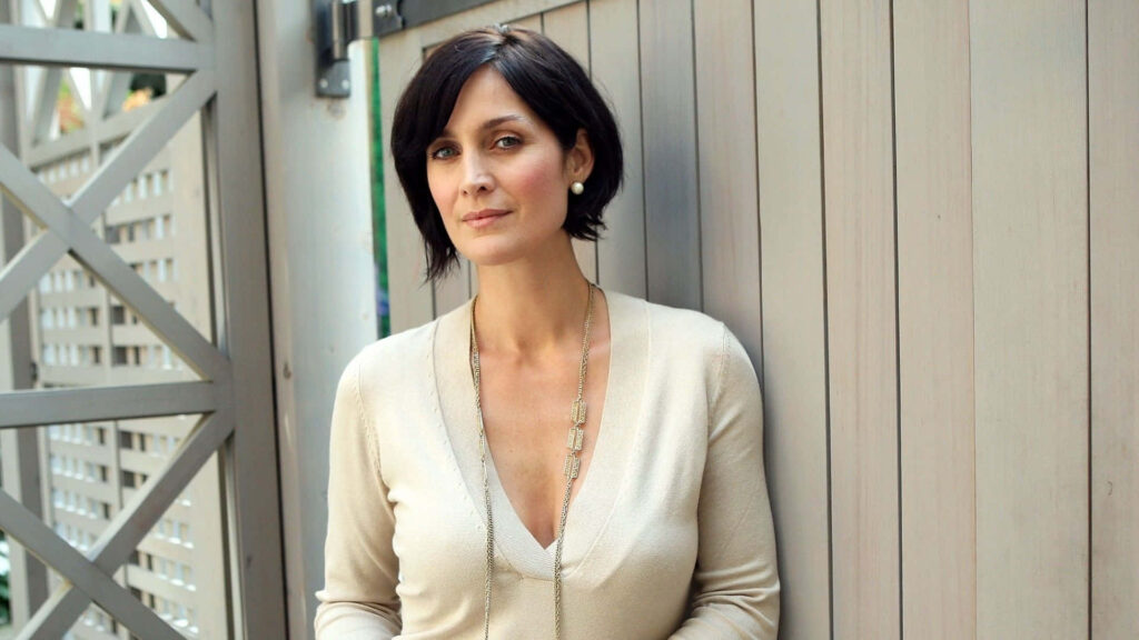 Carrie-Anne Moss in elegant cream outfit against modern wooden fence with geometric patterns background Wallpaper