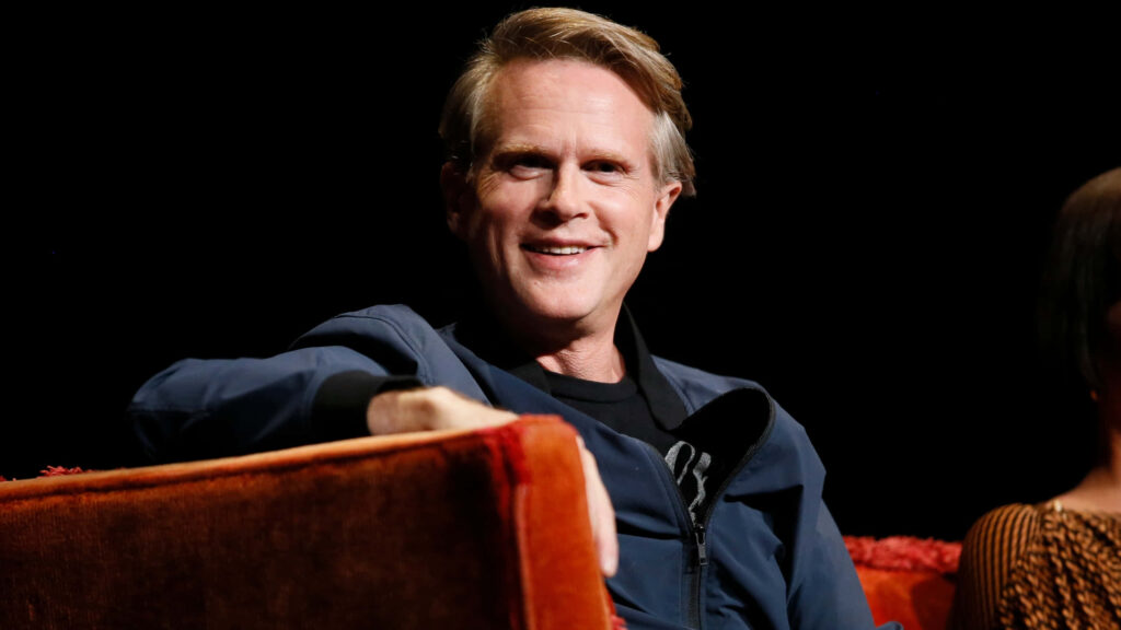 Cary Elwes seated on rust couch with warm smile dark background wallpaper background