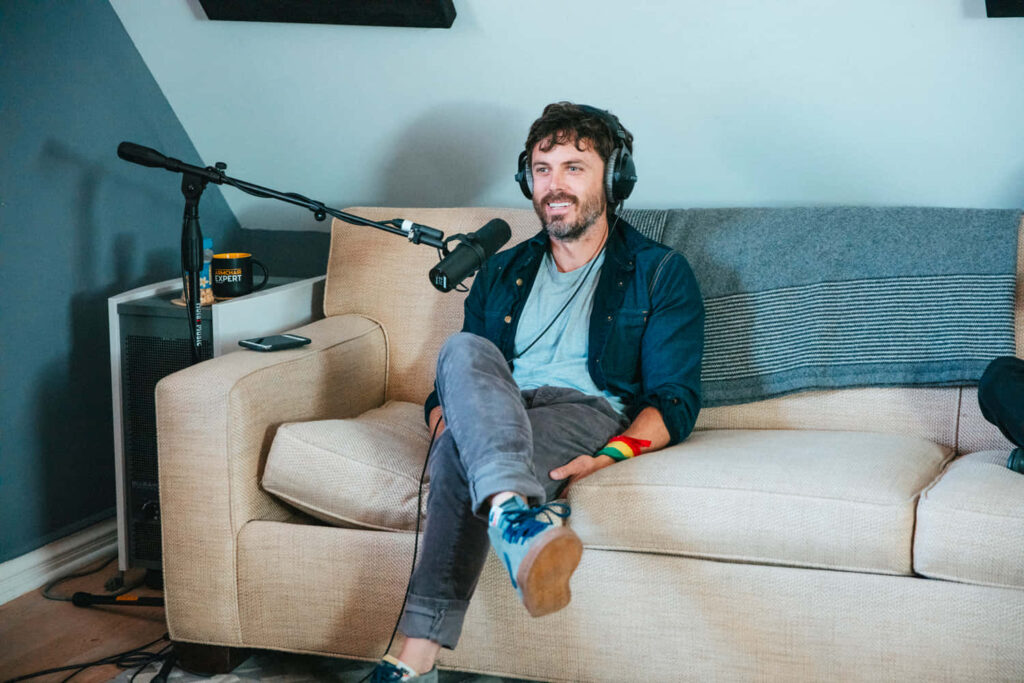Casey Affleck on beige couch podcasting with headphones and microphone in cozy modern studio Wallpaper