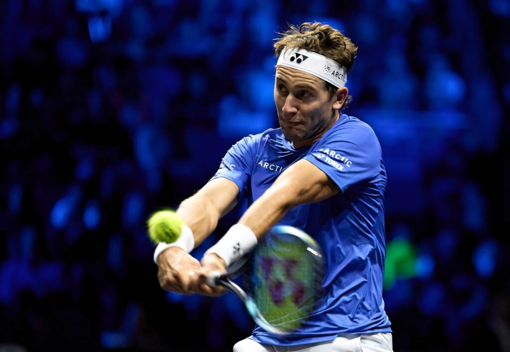 Casper Ruud hitting two handed backhand on blue tennis court under dramatic spotlights action shot Wallpaper