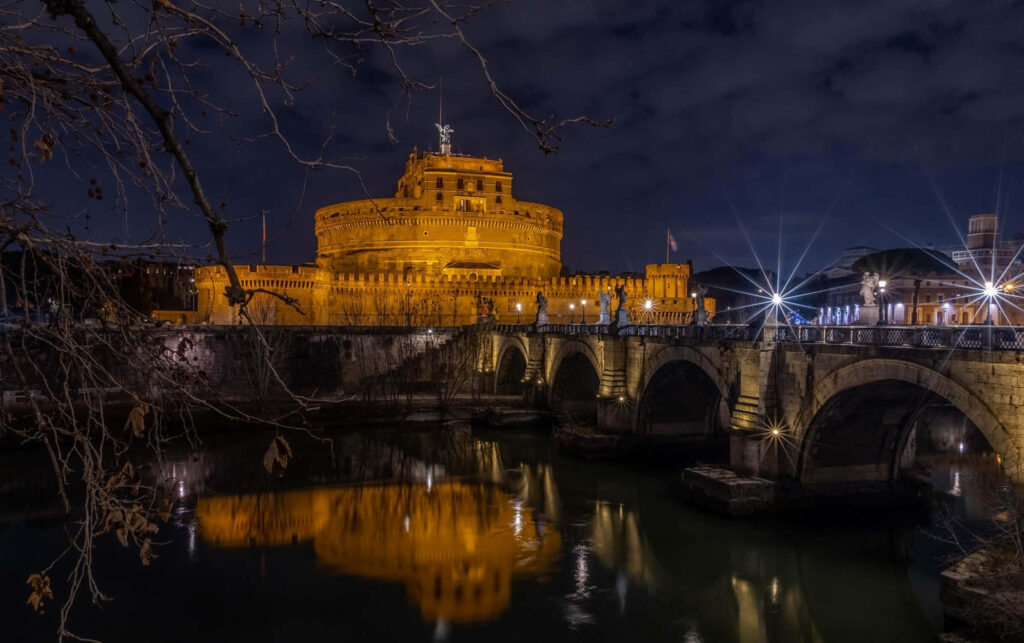 Castel Sant Angelo illuminated at night reflecting on Tiber River with stone bridge background Wallpaper
